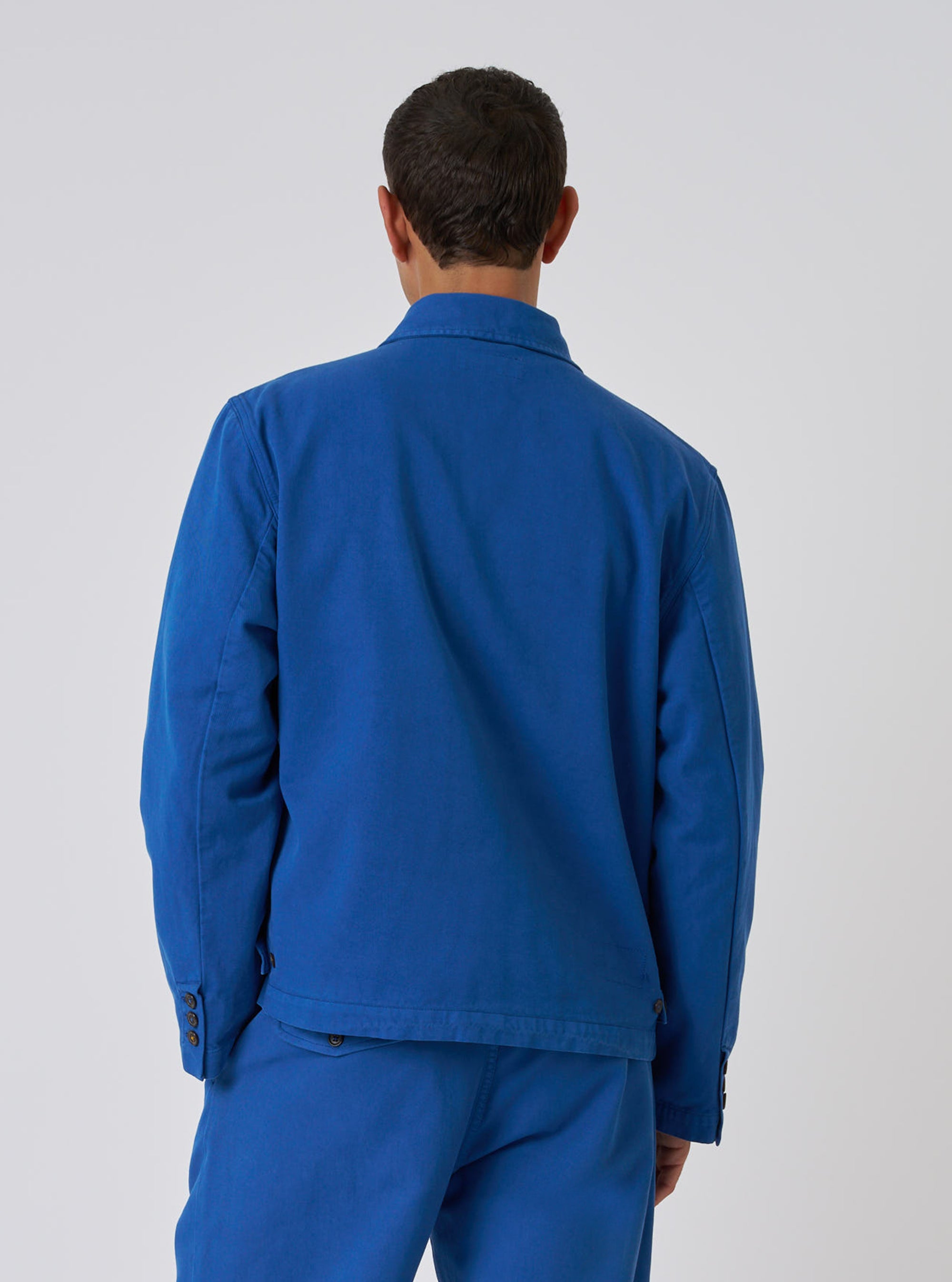 Back view of a model wearing the navy Derek Twill Warmus Jacket. The jacket is a vibrant blue, made of twill, and has a relaxed fit. Shot against a plain white background.