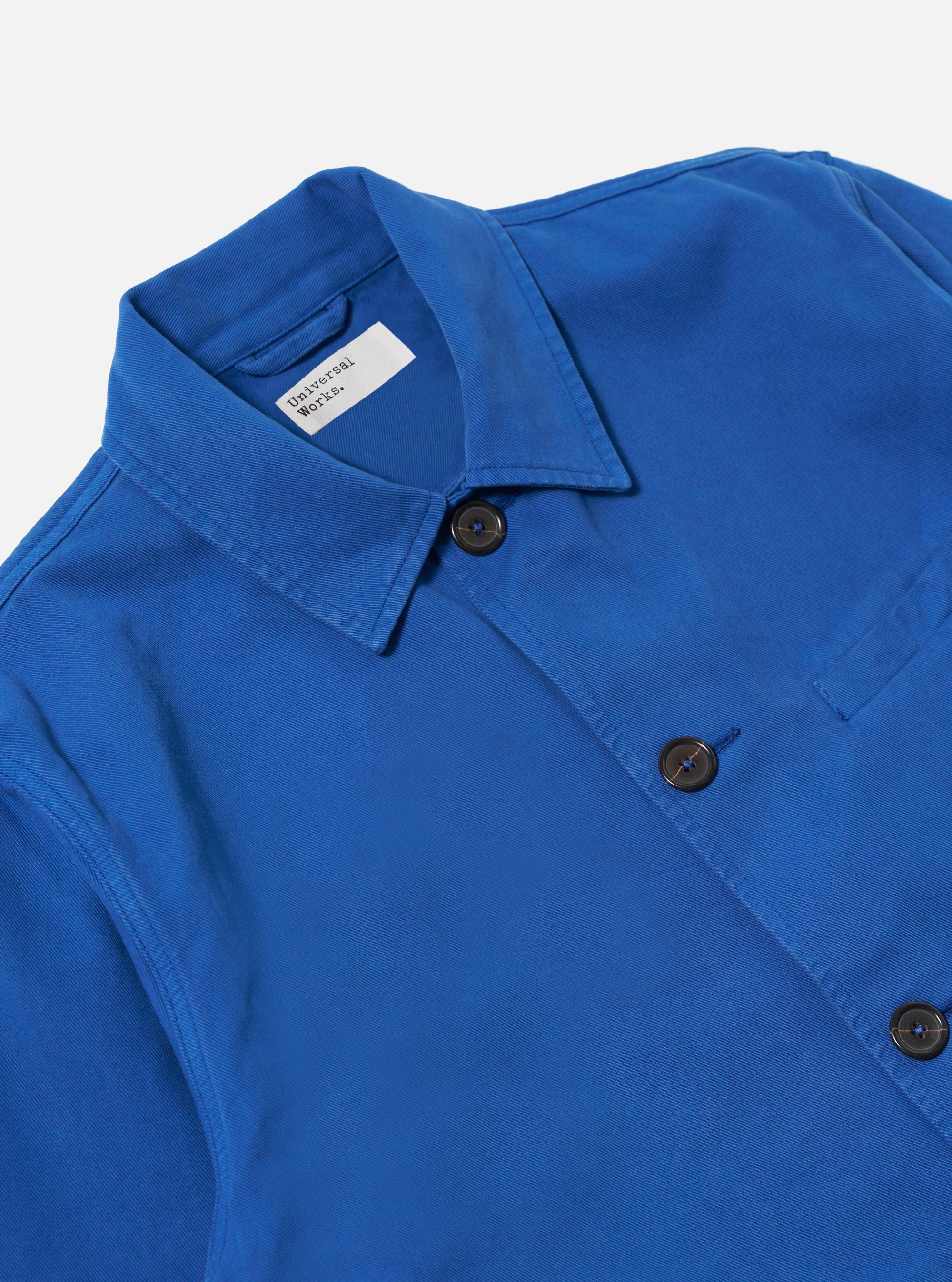 Close-up of the Navy Derek Twill Warmus Jacket on a white background. The jacket is a vibrant blue with brown buttons and a visible brand tag.