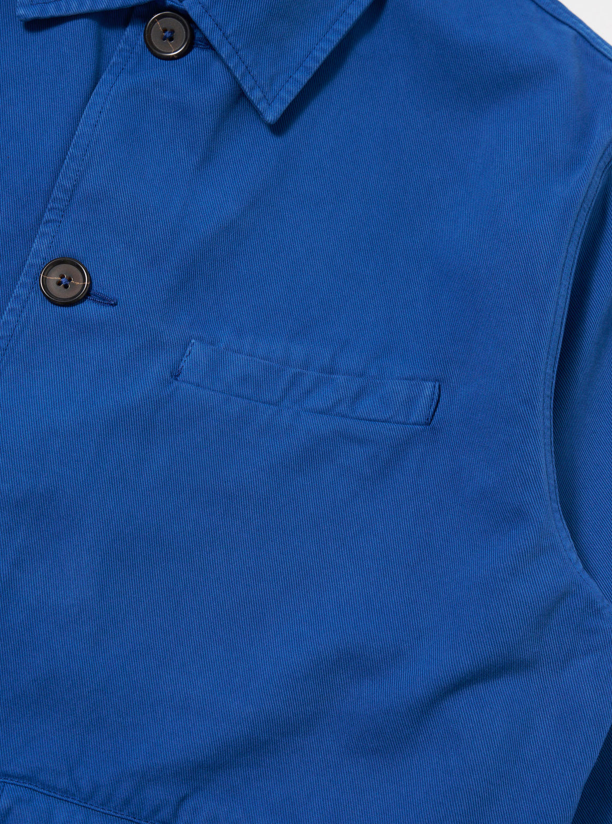 Close-up of the Navy Derek Twill Warmus Jacket. The jacket is a vibrant blue twill with brown buttons. A small pocket is visible. The background is a plain white.