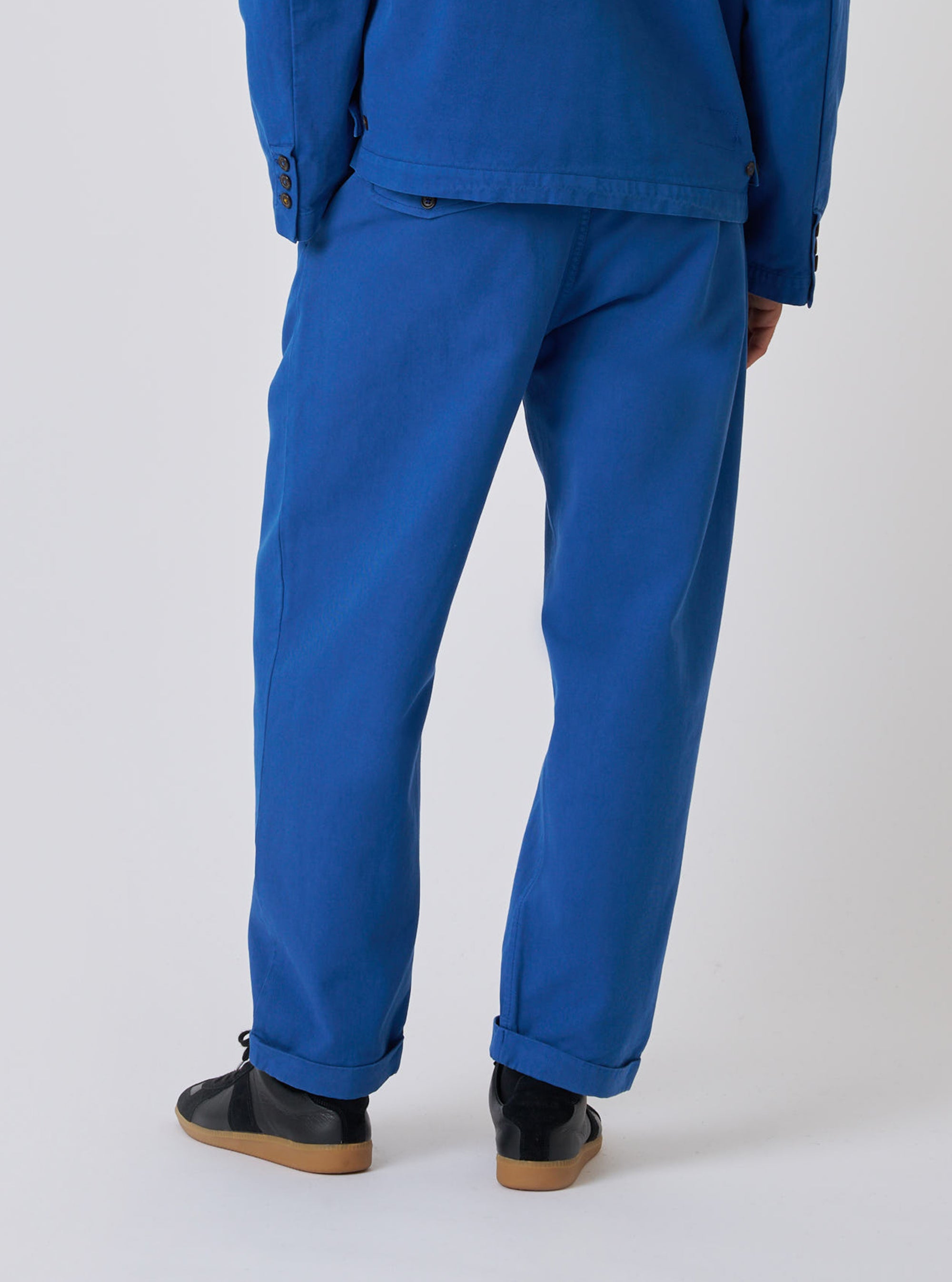 Rear view of a model wearing the Navy Derek Twill Super Chino, a blue twill pant, paired with black sneakers. Shot in a bright studio setting.