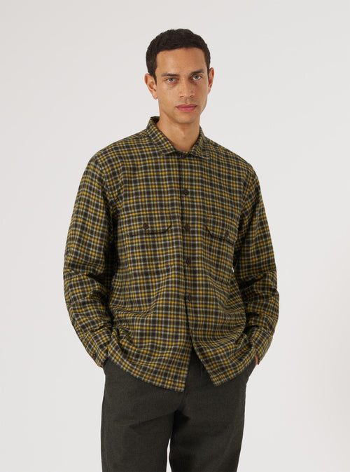 A model poses in front of a white background wearing a Brown Bang Check Utility Shirt. The shirt is a brown and yellow plaid with two chest pockets.