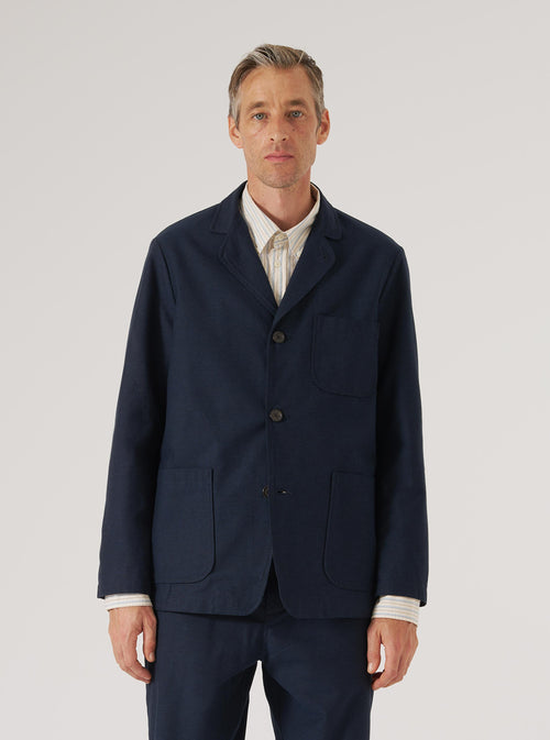 A model poses in a Navy Lucca Suiting Three Button Jacket against a white backdrop. The jacket is a dark navy with three buttons and patch pockets.