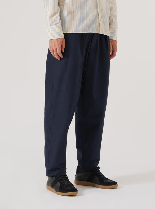 A model on a white background wears the Navy Lucca Suiting Pleated Track Pant. The pants are paired with a striped shirt and black sneakers. The pants have a relaxed fit.