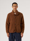 A man stands against a white backdrop, modeling the Cumin Alvar Fleece Lancaster Jacket. The brown fleece jacket features a button-up design and trim.