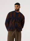 A man poses in front of a white background, wearing a Brown/Navy Argyll Fleece Cardigan. The cardigan has brown and navy diamond patterns with orange lines.