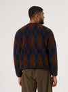 A man stands against a white backdrop, modeling the Brown/Navy Argyll Fleece Cardigan. The cardigan features a brown and navy argyle pattern.