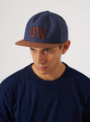 A man wears a Navy/Brown Canvas Baseball Hat in a studio shot. The hat has brown accents and a 'UW' logo. The background is a simple white backdrop.
