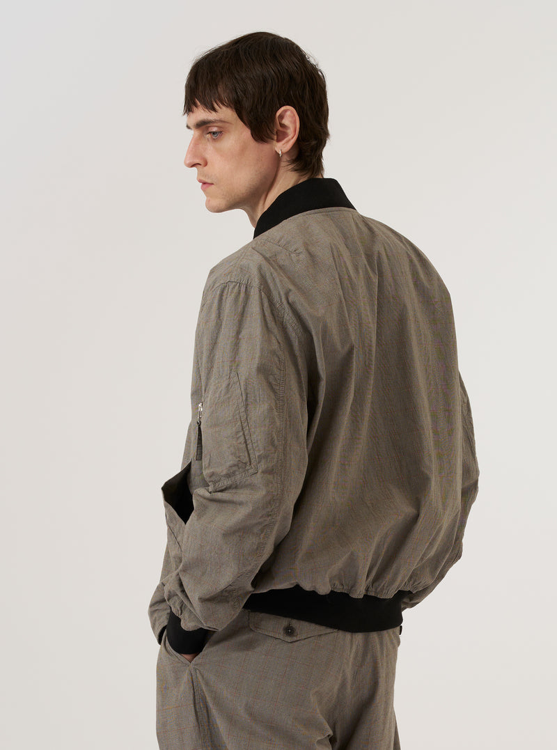 The Grey Brown Wales Glen Check NS Bomber Jacket is worn by a model against a plain white background. The image, captured from the rear, highlights the jacket's relaxed fit and the intricate grey and brown glen check pattern. The garment features a ribbed collar, cuffs, and hem, with a clean, straight-cut back panel. No mannequin is present in the image.