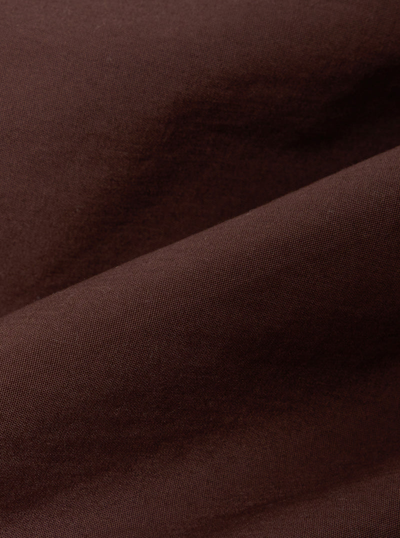 A close-up fabric swatch of the Brown Peached Poplin NS Bomber Jacket highlights the smooth, matte texture and rich brown color of the peached poplin material. The fabric fills the frame, providing a detailed view of the garment's weave and solid color.