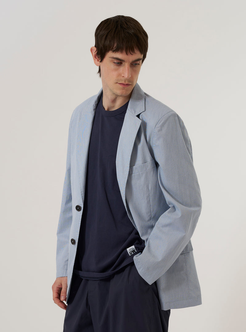 A model wears the Navy Kennedy Stripe Boston Blazer over a light-colored shirt, standing against a plain, neutral background. The blazer features notched lapels, a three-button front closure, and three patch pockets on the front. The fabric is a navy blue cotton with a fine white pinstripe pattern throughout.