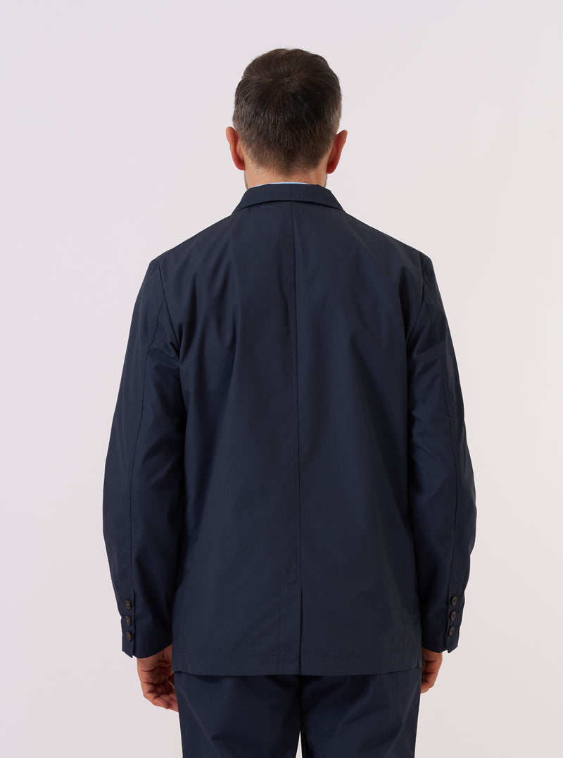 A model wears the Navy Recycled Polytech Capitol Jacket over a light-colored top, shown from the back against a plain white background. The jacket features a clean, structured silhouette with a central seam and a small brand tag at the side. The model is standing with their arms at their sides.