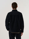 A model wears the Black Organic Fine Cord Duty Jacket over a light-colored top, shown from a rear three-quarter angle against a plain white background. The image highlights the jacket's structured back with a central vertical seam, a straight hem, and the fine-ribbed texture of the black organic cotton corduroy fabric. The sleeve and side profile are also visible.