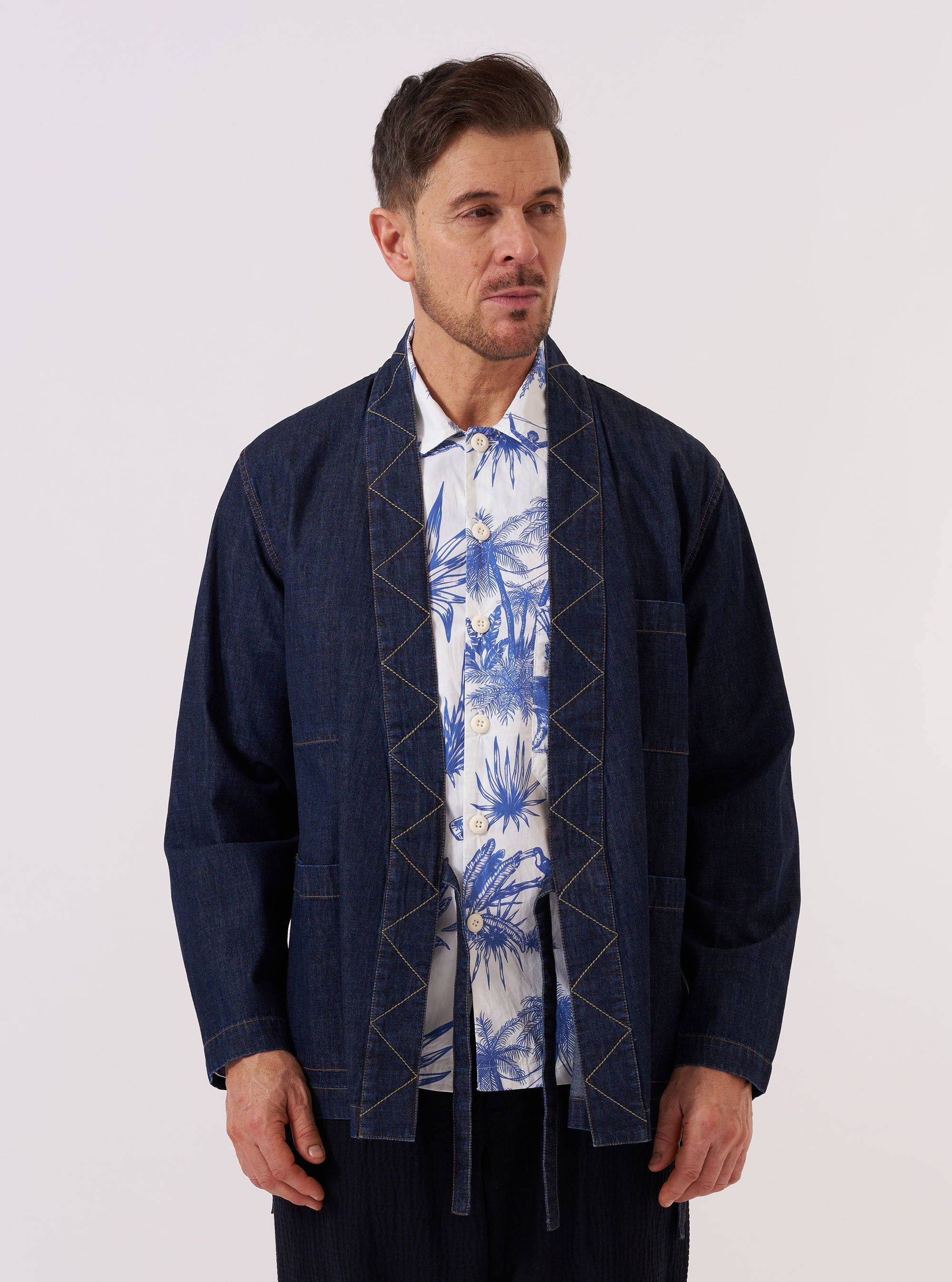 A model wears the Indigo Kaihara LT WT Denim Kyoto Work Jacket against a plain white background. The jacket is a wrap-style, collarless denim top with fabric ties at the side. It features a prominent external chest patch pocket and two lower patch pockets. The model wears the jacket over a light-colored top, and the indigo-washed fabric shows a slight texture.