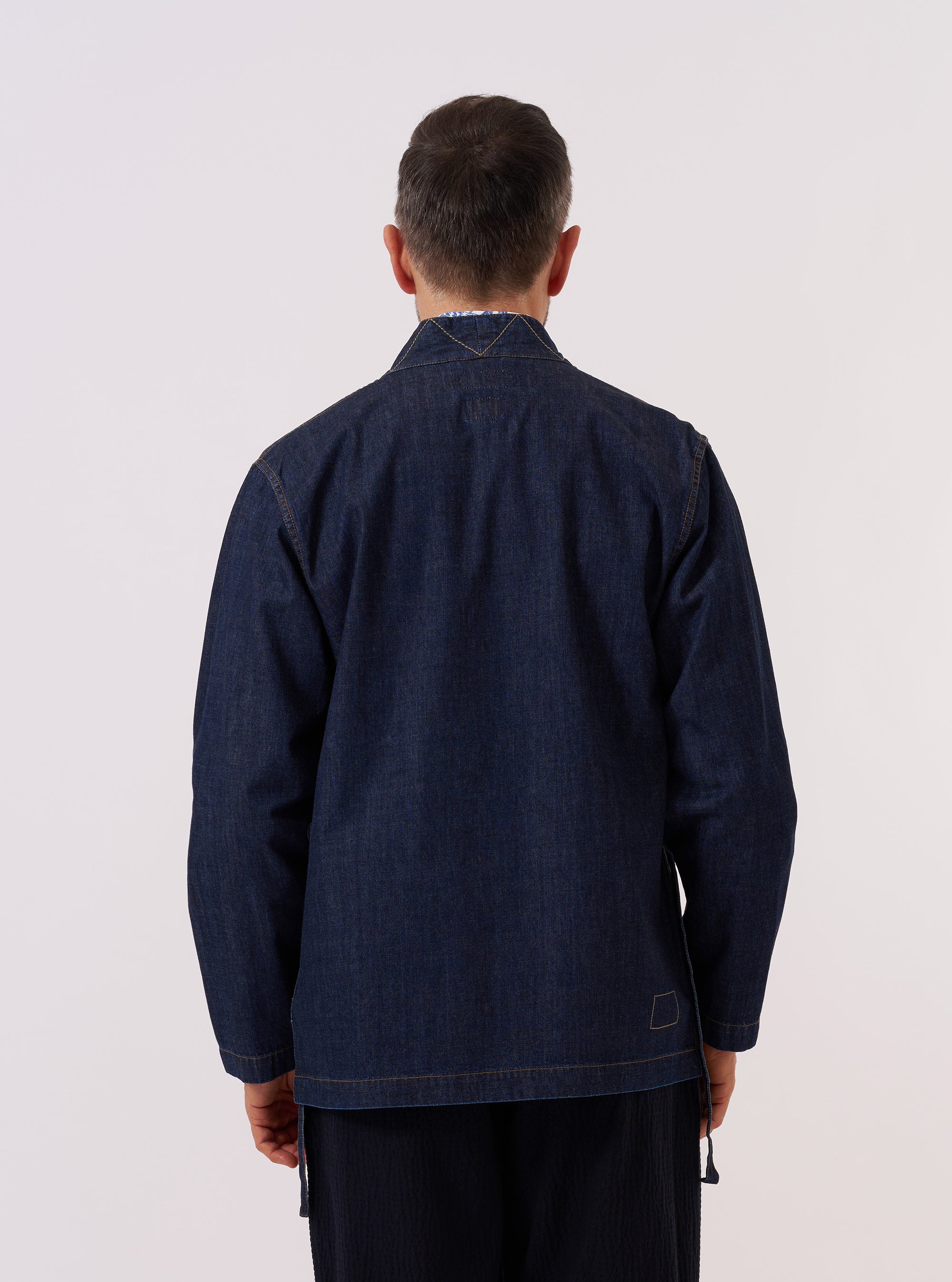A model wears the Indigo Kaihara LT WT Denim Kyoto Work Jacket over a light-colored top, shown from a rear three-quarter angle against a plain white background. The jacket features a wrap-style front with an external tie closure on the side and a straight hem. The deep indigo denim fabric has a visible texture and a relaxed fit through the sleeves and body.