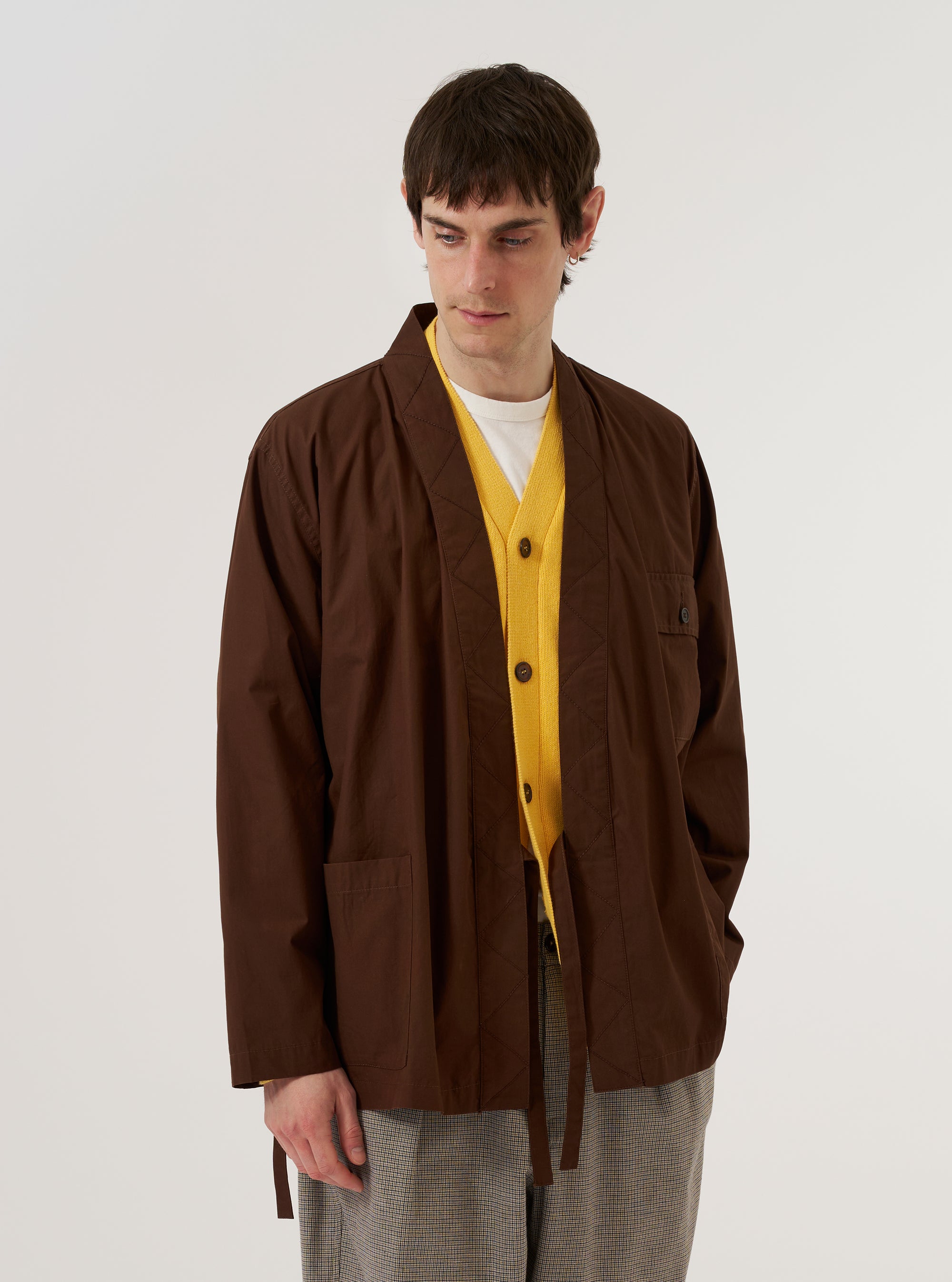 A model wears the Brown Peached Poplin Osaka Work Jacket against a plain white background. The jacket features a wrap front with internal and external tie closures, a chest patch pocket with a button flap, and military-inspired contrast embroidery. The medium-weight brown cotton fabric has a soft, peached finish. The model layers the jacket over a light-colored top.