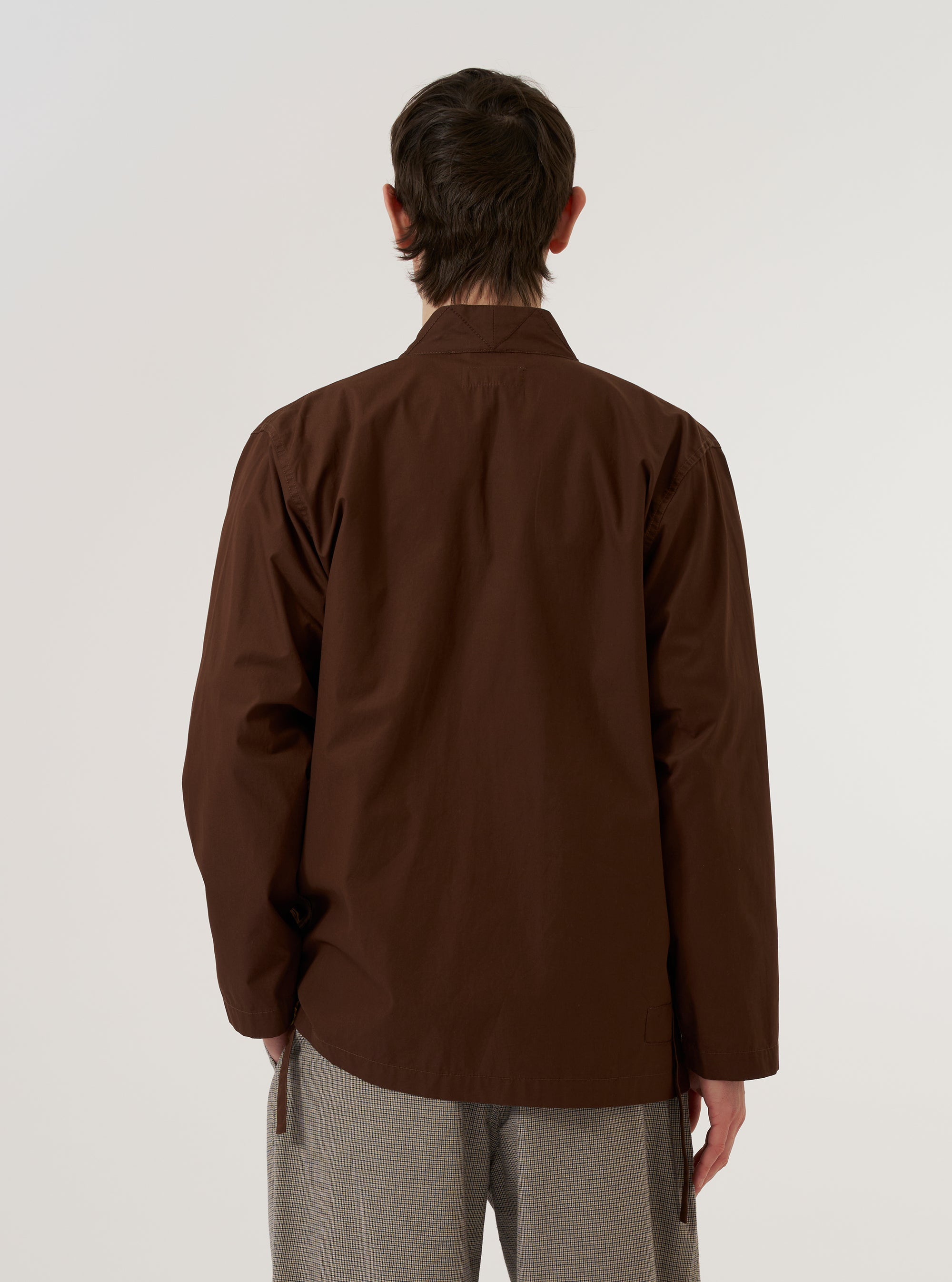 A model wears the Brown Peached Poplin Osaka Work Jacket over a light-colored shirt, shown from a rear three-quarter angle against a plain, neutral background. The jacket features a relaxed, wrap-style design with wide sleeves and a straight hem. The fabric is a solid brown peached poplin with a smooth, matte finish.