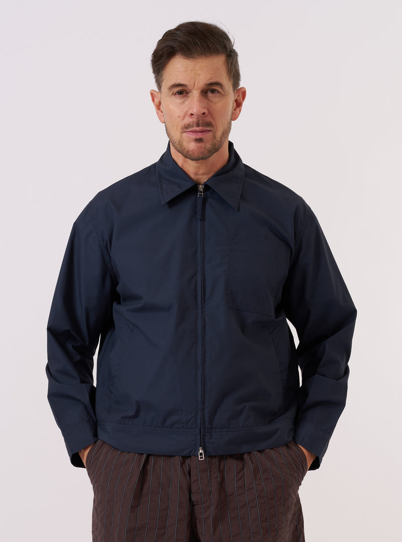 A model wears the Navy Recycled Polytech Rose Bowl Jacket over a light-colored shirt, standing against a plain, neutral studio background. The jacket features a short, boxy fit with a central two-way zip closure and a slightly oversized collar. Prominent details include a large patch pocket on the left chest and two slanted welt pockets at the waist. The smooth, dark navy fabric has a clean, utilitarian appearance. No mannequin is present in the image.