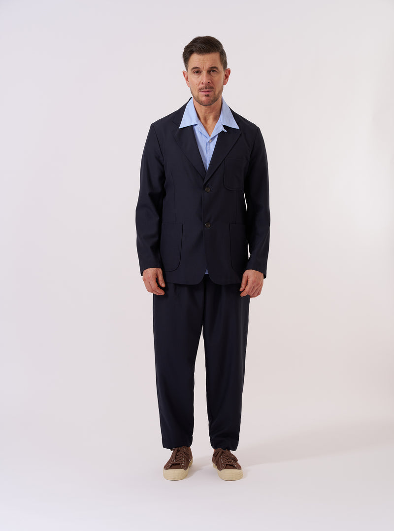 The Navy Tropical Suiting Two Button Jacket is displayed as a flat lay against a plain white background. The jacket features a relaxed fit with a two-button front closure, a wide notch lapel, and three front patch pockets, including one at the chest and two at the waist. The navy blue fabric is a lightweight tropical wool blend with a smooth, refined texture.
