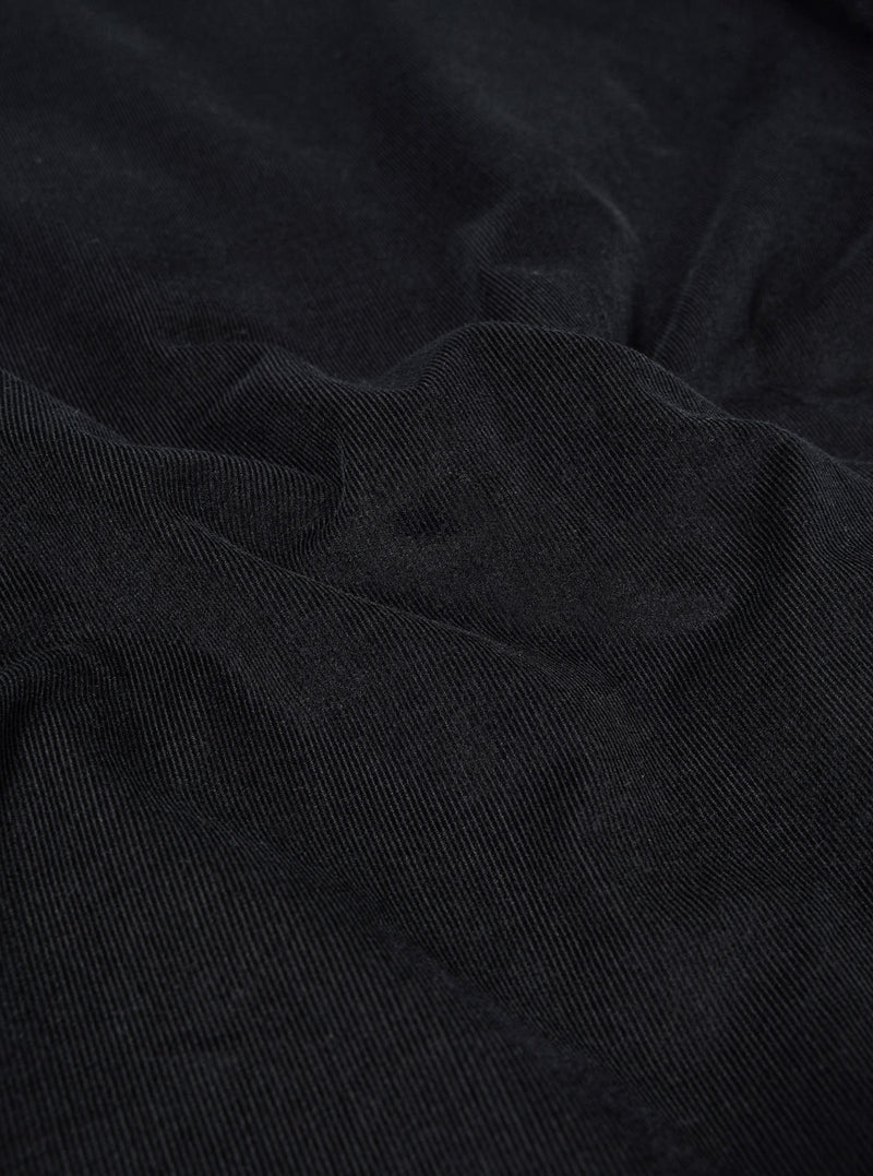 A close-up swatch of the Black Organic Fine Cord Two Button Jacket highlights the fine-wale texture and vertical ribbed pattern of the black organic cotton corduroy fabric. The material fills the frame, providing a detailed view of the garment's color and weave.