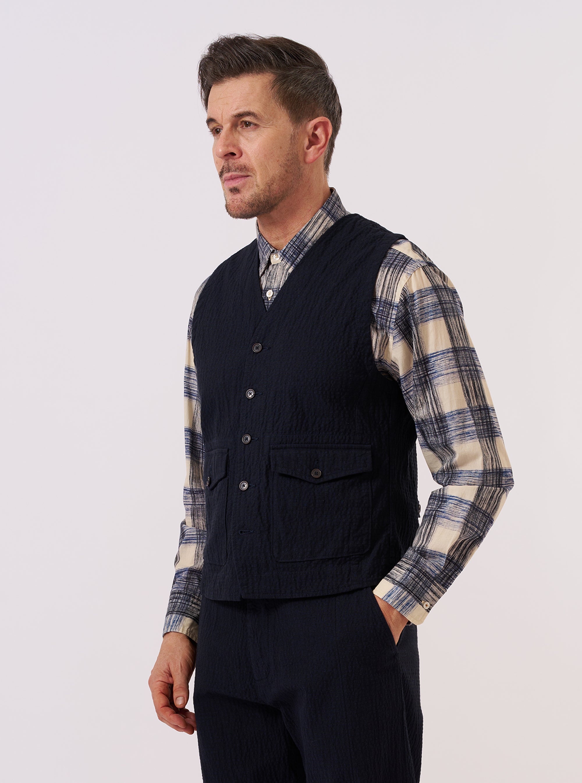 A model wears the Dark Navy Ospina Cotton Field Waistcoat over a light-colored shirt, standing against a plain, neutral background. The waistcoat features four large front patch pockets, a structured collar, and a central button closure. The dark navy fabric has a visible seersucker texture.