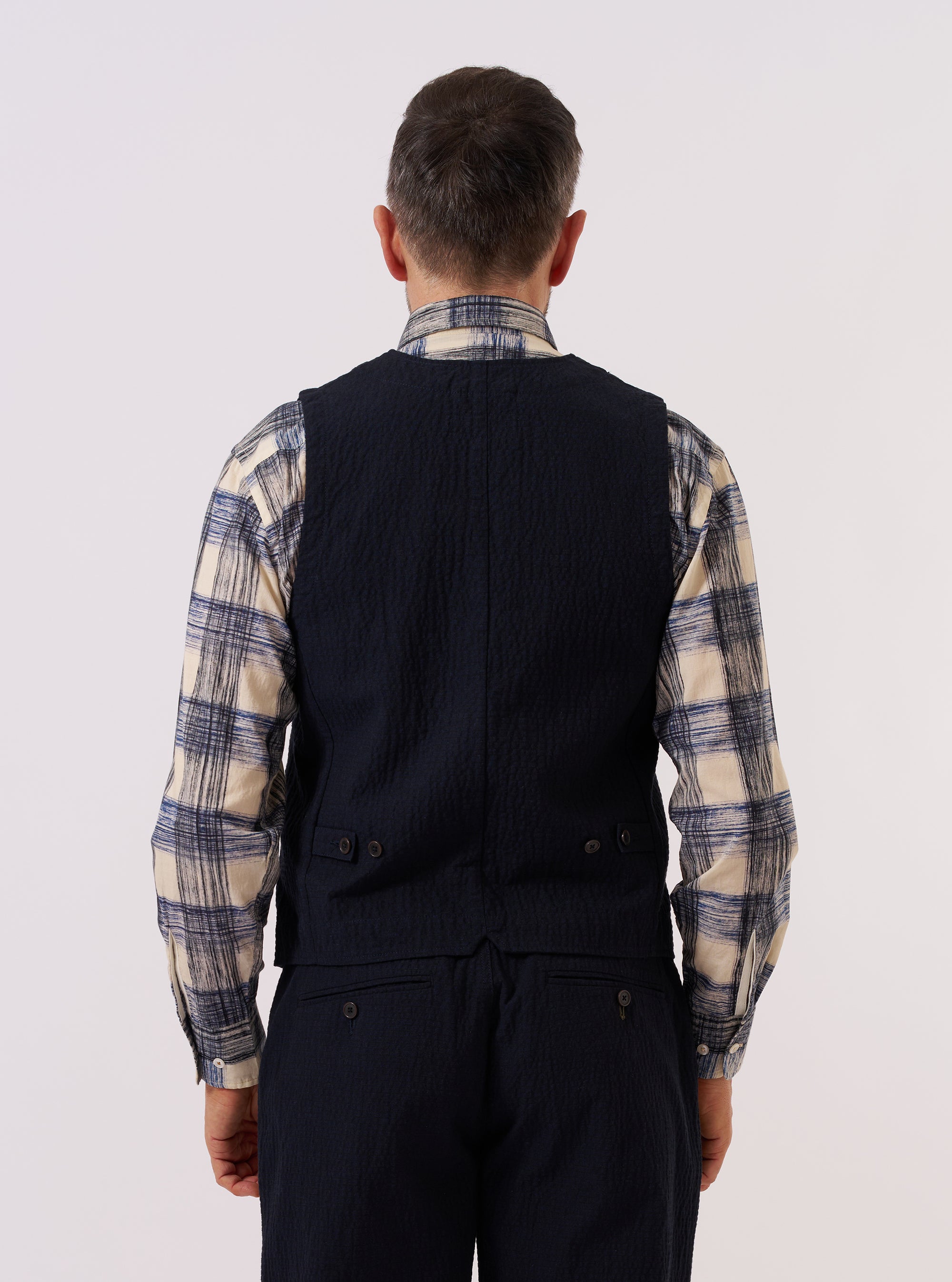 A model wears the Dark Navy Ospina Cotton Field Waistcoat over a light-colored shirt, shown from a rear three-quarter angle against a plain, neutral studio background. The waistcoat features a structured back with vertical seams and a straight hem. The dark navy fabric has a subtle seersucker texture.