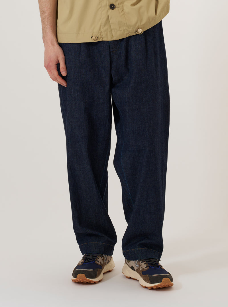 The Indigo Kaihara LT WT Denim Duke Pant is worn by a model against a plain, light-colored background. The image shows a front view of the trousers, highlighting the wide-leg fit, single pleat at the waist, and a central button fly. The model is wearing the indigo blue denim pants with a white t-shirt tucked in, showcasing the fixed waistband and relaxed silhouette.
