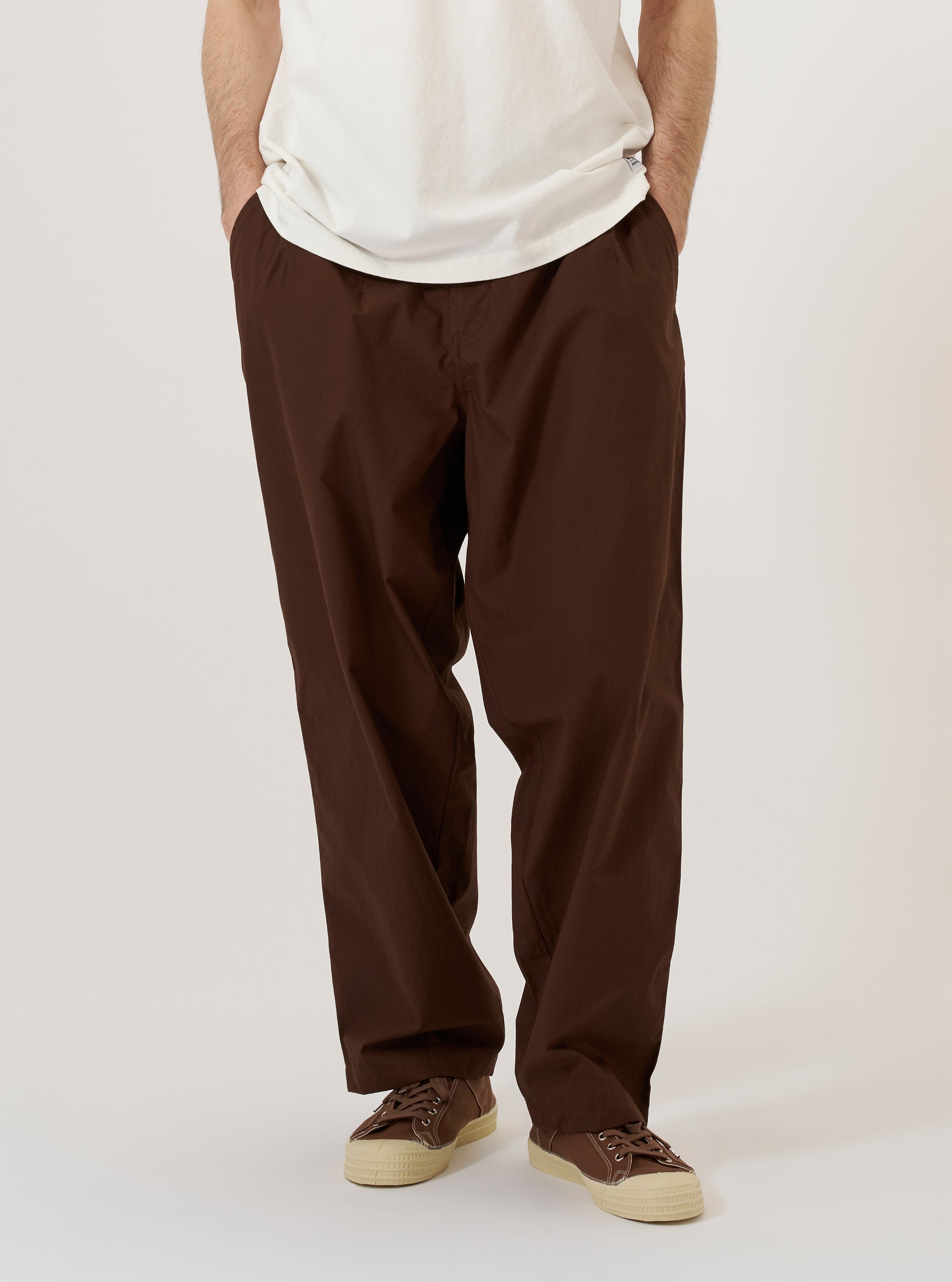 A model is wearing the Brown Peached Poplin Dutch Pant while standing against a plain white background. The trousers feature a loose, wide-leg fit and are shown from the front, paired with a light-colored shirt and shoes. The peached poplin fabric has a smooth, matte finish.