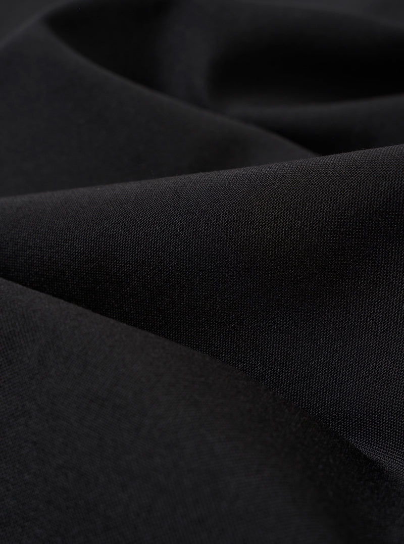 A close-up fabric swatch of the Black Tropical Suiting Military Chino highlights the smooth, technical texture and deep black color of the polyester and wool blend material. The fabric fills the entire frame, showcasing the fine weave of the tropical suiting.