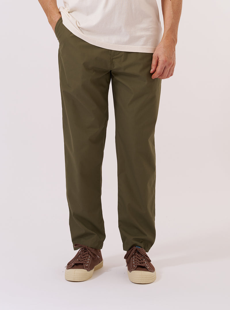 A model is wearing the Olive Recycled Polytech Military Chino, shown from the waist down against a plain white background. The olive green trousers feature a contemporary slim-straight fit, a button fly, and two side pockets. The model is wearing a light-colored top and white socks with black shoes to highlight the clean lines and tapered leg of the chinos.