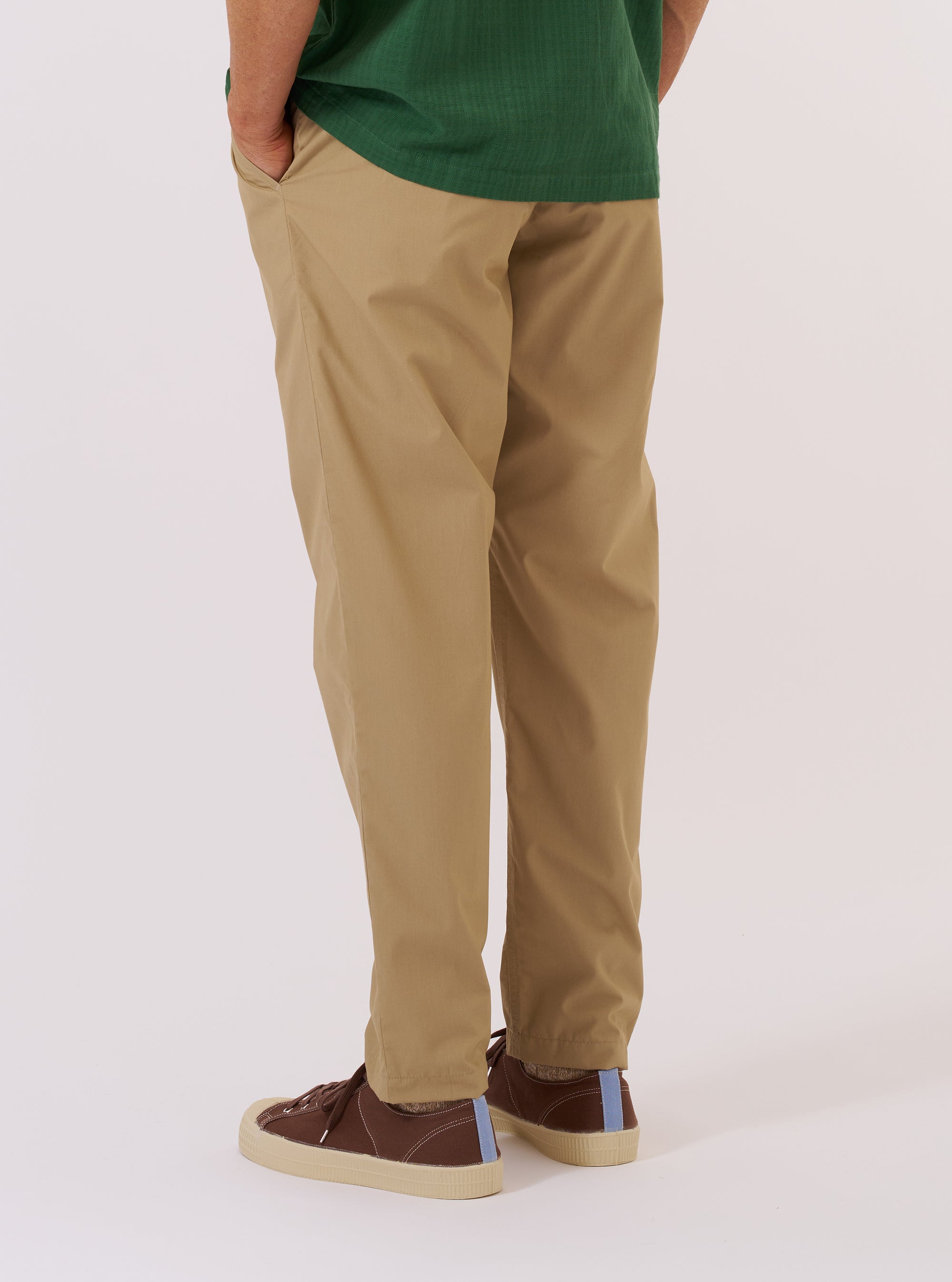 A model wears the Sand Recycled Polytech Military Chino, shown from a rear angle against a plain white background. The image highlights the slim, straight-leg fit and the clean construction of the back, including two rear welt pockets. The trousers are made from a smooth, sand-colored recycled polytech fabric.