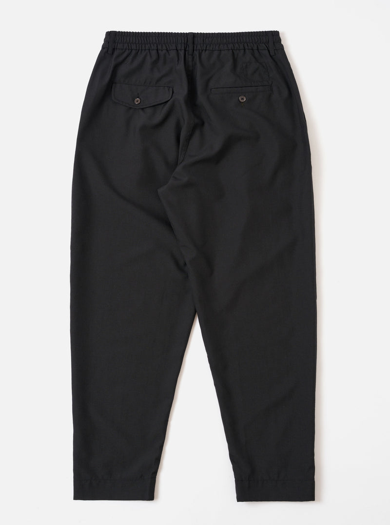 The Black Tropical Suiting Pleated Track Pant is shown from the back as a flat lay against a plain white background. The image highlights the clean construction of the trousers, featuring two rear pockets: one buttoned flap pocket on the left and one button jetted pocket on the right. The black tropical suiting fabric appears smooth and lightweight.