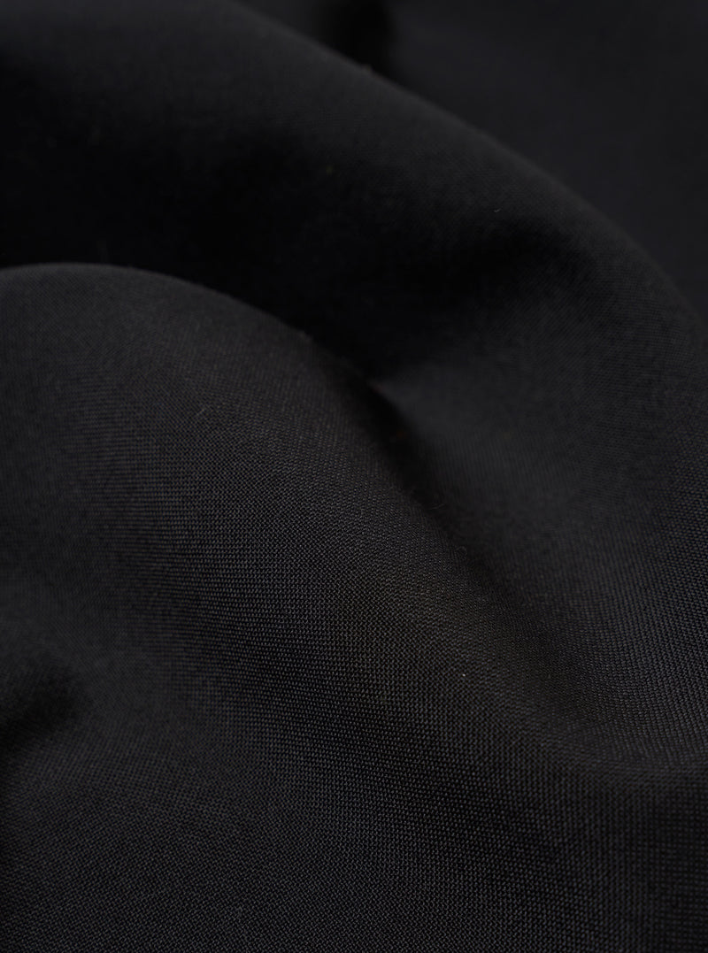 A close-up fabric swatch of the Black Tropical Suiting Pleated Track Pant is shown, highlighting the fine, smooth texture and solid black color of the lightweight tropical suiting material.