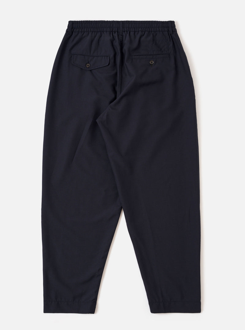 The Navy Tropical Suiting Pleated Track Pant is shown from the back as a flat lay against a plain white background. The image highlights the elasticated waistband and the two rear pockets, one of which features a buttoned flap. The navy wool-blend fabric has a smooth, clean finish.