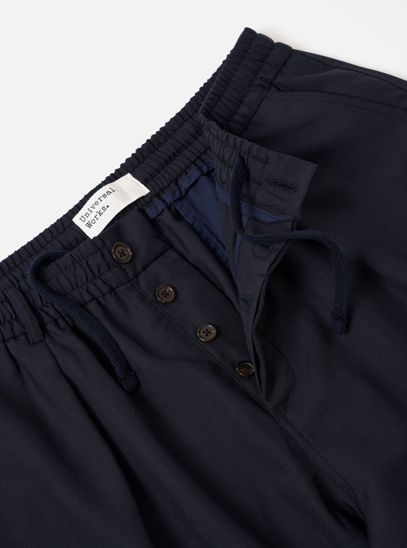The Navy Tropical Suiting Pleated Track Pant is shown from the back as a flat lay against a plain white background. The image highlights the two rear set-in pockets, one with a buttoned flap and one button-jetted, as well as the elasticated waistband. The fabric is a smooth, dark navy tropical suiting with a visible fine-weave texture.