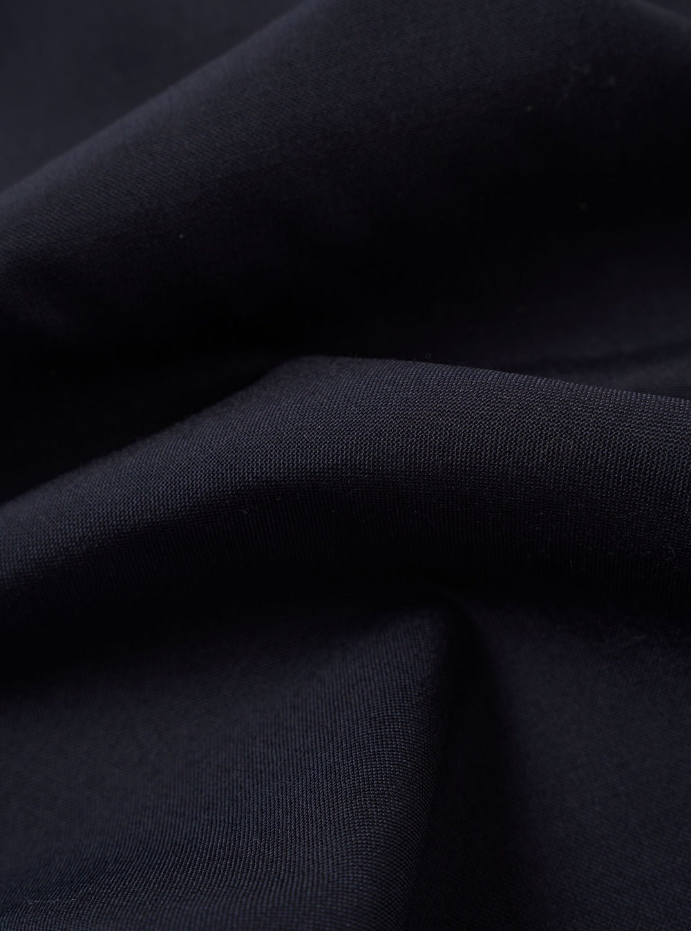 A close-up fabric swatch of the Navy Tropical Suiting Pleated Track Pant highlights the smooth, fine weave and deep navy blue color of the polyester and wool blend material. The fabric fills the frame, showcasing its lightweight and crease-resistant texture.