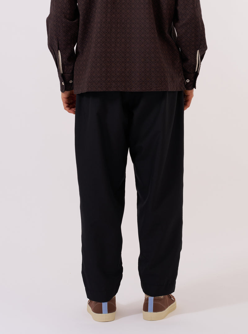 A model is wearing the Black Tropical Suiting Pleated Track Pant, shown from a rear three-quarter angle against a plain white background. The image highlights the relaxed fit and the clean, smooth texture of the black tropical suiting fabric. The trousers feature an elasticated waistband and a distinct taper toward the hem.