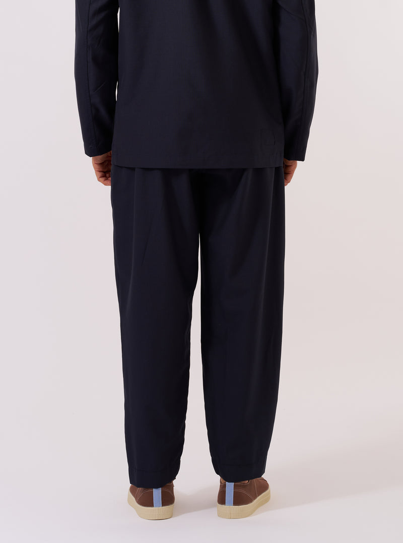 A model is wearing the Navy Tropical Suiting Pleated Track Pant, shown from the waist down against a plain white background. The image highlights the loose, tapered fit of the trousers and the single pleat at the front. The model is wearing the pants with a white t-shirt tucked into the elasticated waistband and white socks. The navy blue fabric appears smooth and lightweight.
