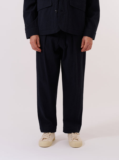 A model wears the Dark Navy Ospina Cotton Oxford Pant as part of a full suit, standing against a plain, neutral background. The trousers feature a wide, loose leg with a single pleat at the front and a visible drawstring at the elasticated waistband. The dark navy fabric has a distinct puckered seersucker texture. The model is shown from a front angle, also wearing a matching jacket and a light-colored shirt.