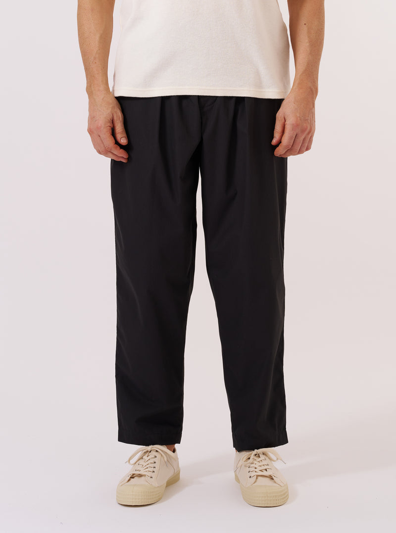 A model wears the Black Recycled Polytech Pleated Track Pant, shown from the waist down against a plain white background. The image highlights the loose, tapered fit and the single pleat at the front. The black trousers feature an elasticated waistband with a drawstring, side pockets, and a button fly. The model is also wearing a white T-shirt and black shoes.