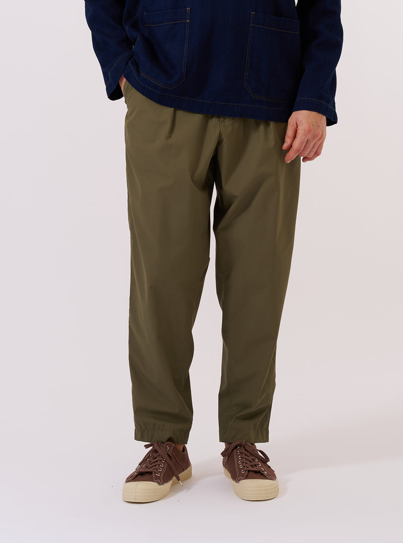 A model wears the Olive Recycled Polytech Pleated Track Pant while standing against a plain white background. The trousers feature a loose fit with a tapered leg, a single pleat at the front, and an elasticated waistband with a drawstring. The model is wearing the pants with a white t-shirt and white socks, highlighting the smooth, olive green technical fabric.