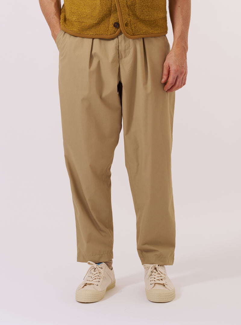 A model wears the Sand Recycled Polytech Pleated Track Pant while standing against a plain white background. The image shows a front view of the trousers, highlighting the loose fit, tapered leg, and the single pleat at the front. The elasticated waistband and the light sand-colored recycled poly-tech fabric are prominent. The model is wearing the trousers with a light-colored top tucked in to display the waistband detail.