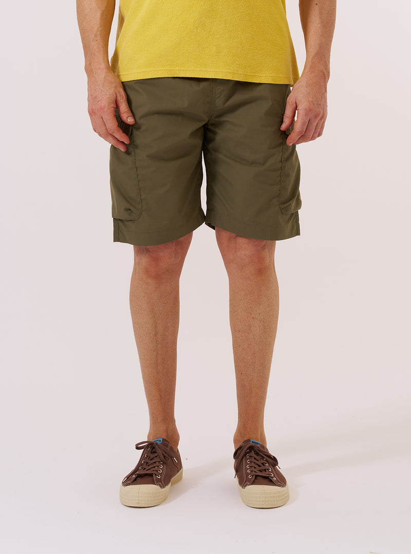 A model wears the Olive Recycled Polytech Parachute Short while standing against a plain white background. The shorts feature a relaxed, wide-leg fit, an elasticated waistband with an integrated drawstring, and large side pockets. The garment is made from a smooth, olive green technical fabric. The model is shown from the waist down, providing a clear view of the front and side profile of the shorts.
