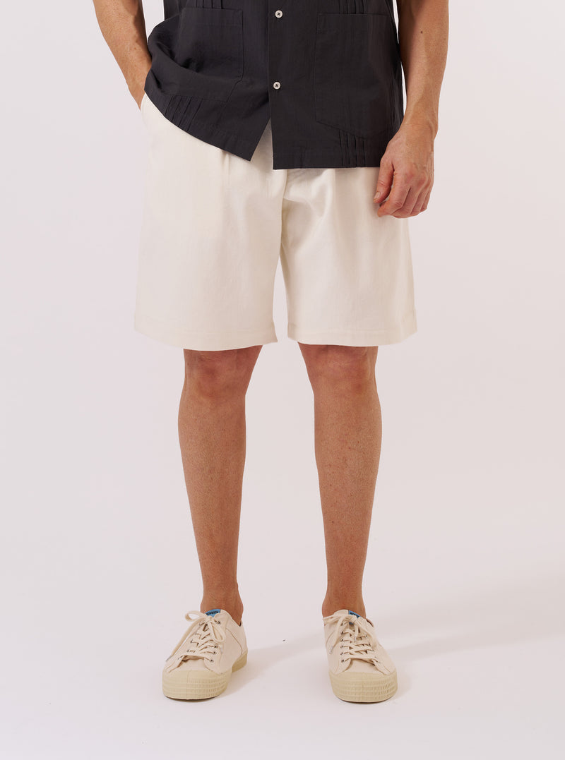 A model wears the Ecru Slub Drill Pleated Track Short, standing against a plain white background. The image shows a front view of the shorts, highlighting the elasticated waistband, the distinct single pleat on each leg, and the textured ecru cotton slub drill fabric. The model is wearing a light-colored shirt tucked into the shorts.