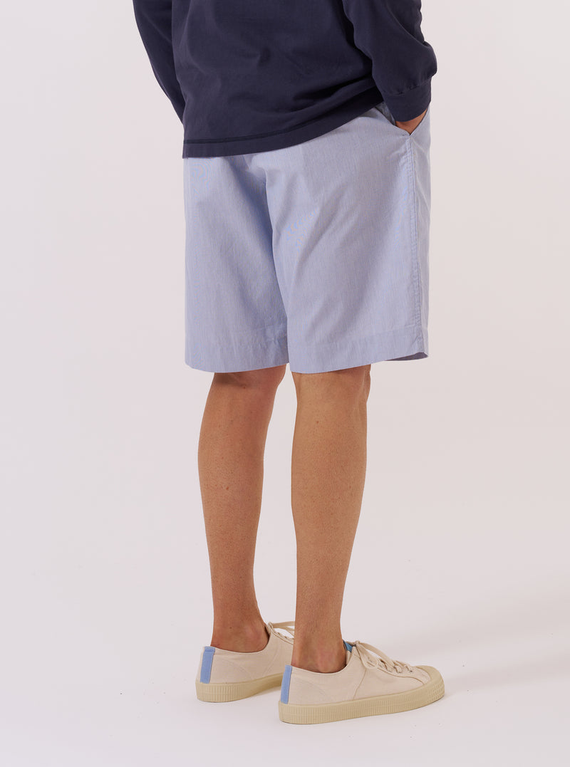 The Navy Kennedy Stripe Pleated Track Short is shown from a rear three-quarter angle as a flat lay against a plain white background. The shorts feature a vertical white pinstripe pattern on a navy blue base, an elasticated waistband, and a single rear patch pocket on the right side. The fabric appears to be a structured cotton.