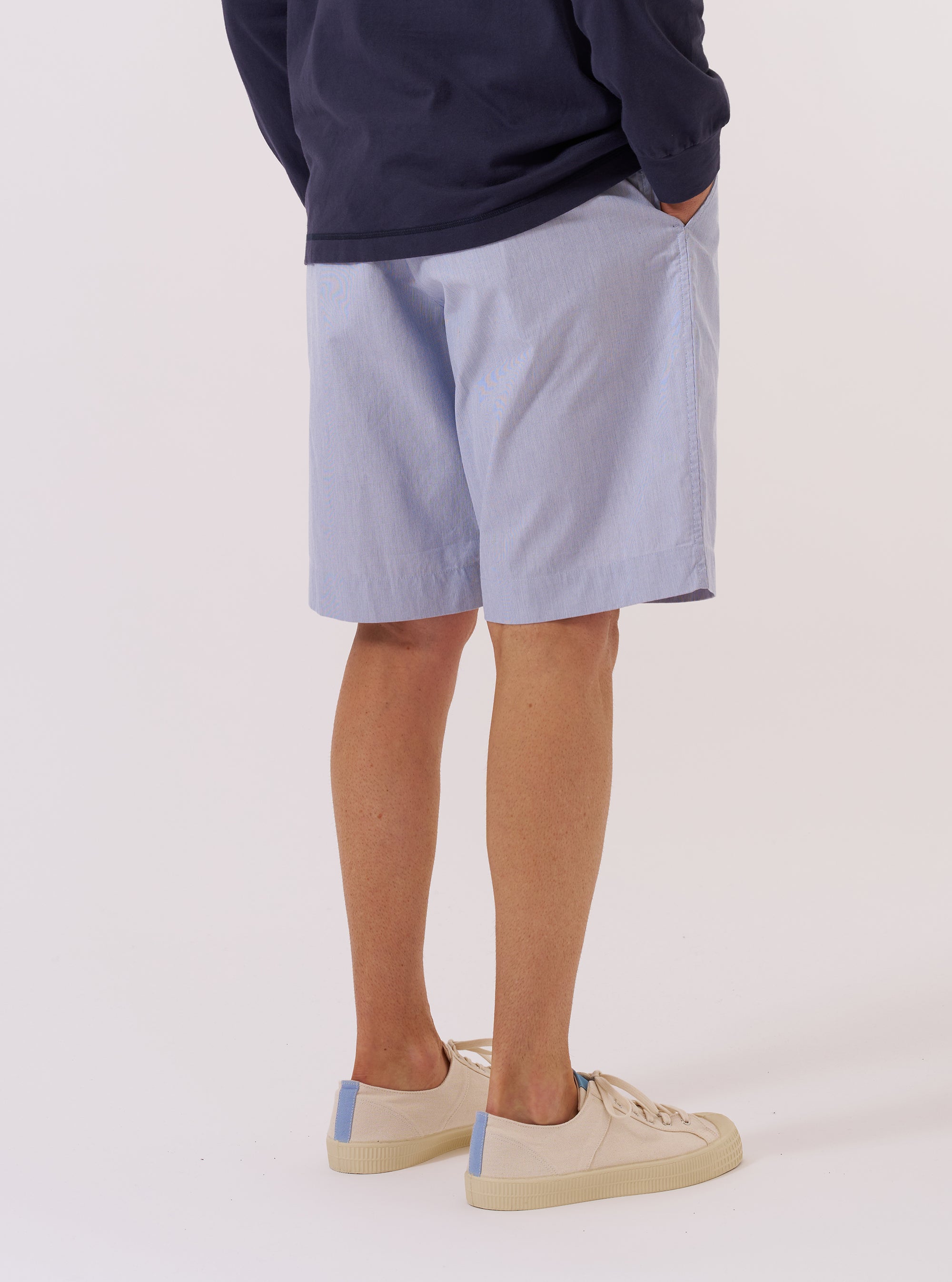 The Navy Kennedy Stripe Pleated Track Short is shown from a rear three-quarter angle as a flat lay against a plain white background. The shorts feature a vertical white pinstripe pattern on a navy blue base, an elasticated waistband, and a single rear patch pocket on the right side. The fabric appears to be a structured cotton.