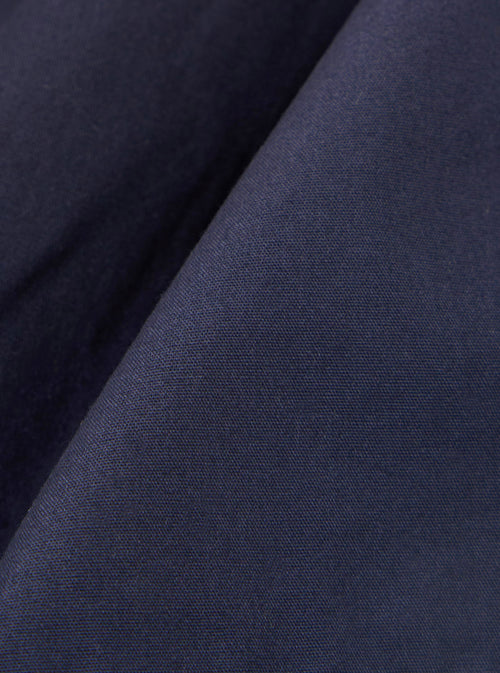 A close-up fabric swatch of the Navy Peached Poplin Pleated Track Short highlights the smooth, matte texture and deep navy blue color of the peached poplin material. The fabric fills the frame, showcasing its fine weave and soft finish.