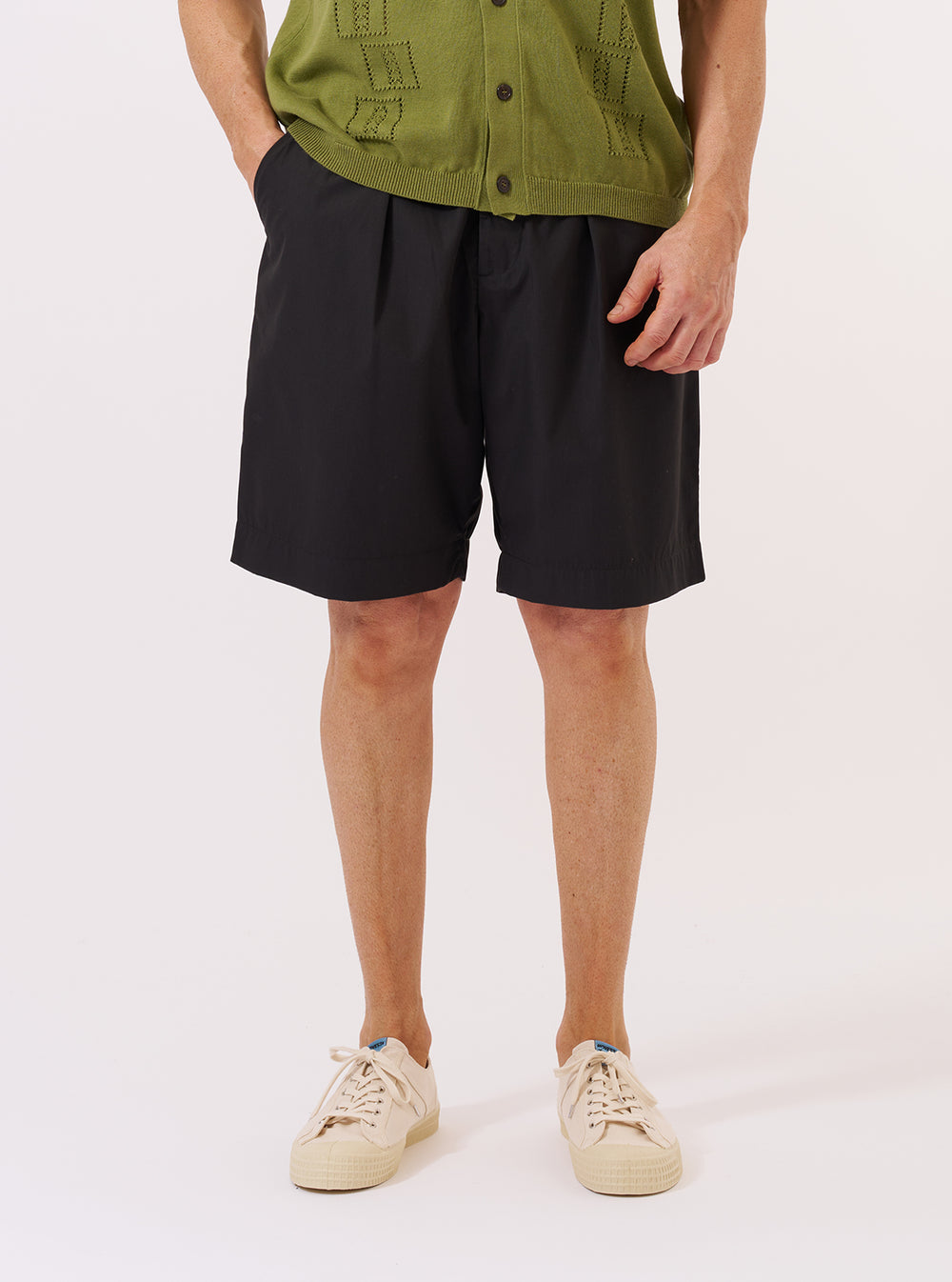 A model wears the Black Recycled Polytech Pleated Track Short against a plain white background. The image shows a front view of the shorts, highlighting the relaxed fit, single front pleats, and an elasticated waistband with a tonal drawstring. The black fabric has a smooth, technical finish. The model is shown from the waist down, wearing a white t-shirt tucked into the waistband.