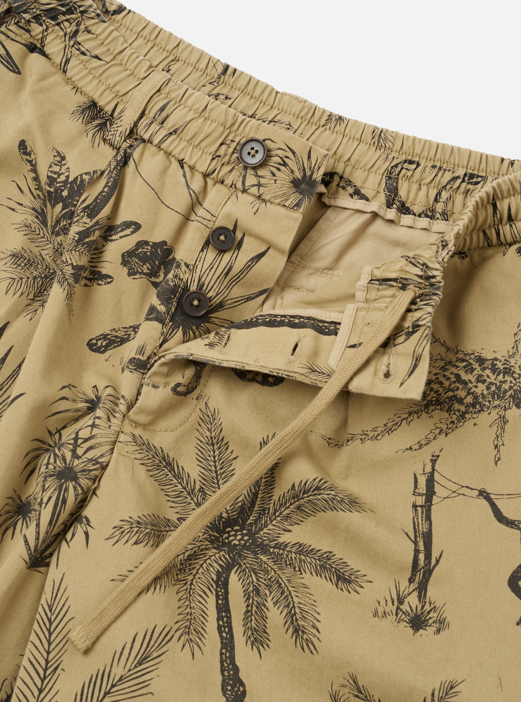 The Sand Jungle Toile Twill Pleated Track Short is shown from the back as a flat lay against a plain white background. The shorts feature a relaxed fit, an elasticated waistband, and two rear patch pockets, with the right pocket featuring a button-down flap. The sand-colored cotton twill fabric is covered in a dark, detailed jungle toile print featuring botanical and animal illustrations.