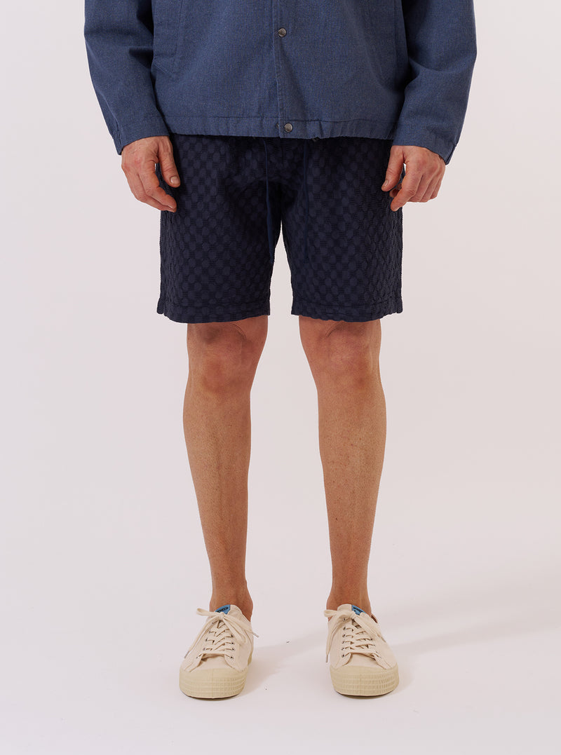 A model wears the Navy Dot Waffle Simple Short while standing against a plain white background. The image shows a front view of the shorts, highlighting the elasticated waistband and the textured dot waffle cotton fabric. The model is wearing the shorts with a white T-shirt tucked in and white socks.