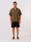The Olive Cape Block Print Camp Shirt is presented as a flat lay against a plain white background. The shirt features a camp collar, a single chest patch pocket, and a front button closure. The fabric is decorated with a repeating block-printed pattern of white and green leaf motifs.